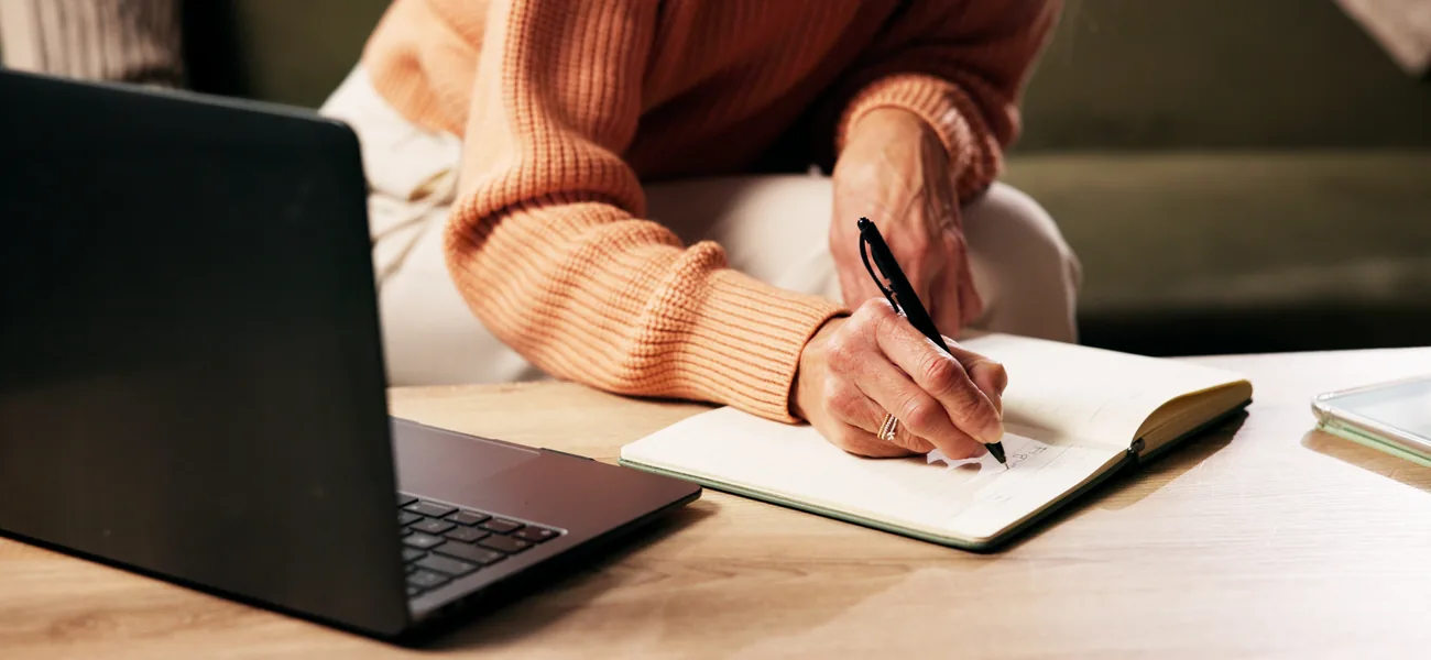 Woman taking notes in a notebook during a telehealth appointment on her laptop